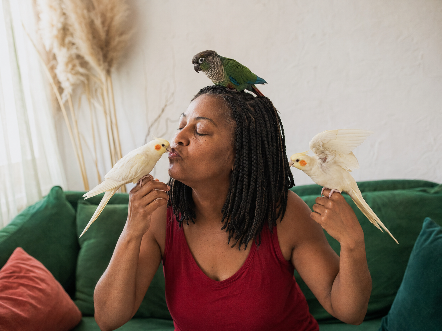 A woman kissing a cockatiel while two more cockatiels perch on her hands and a green conure sits on her head